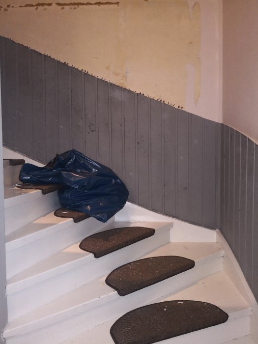 Trappa med vitmålade steg och grå sidopanel som behöver renovering, delvis borttagen tapet och höjdskillnad syns.