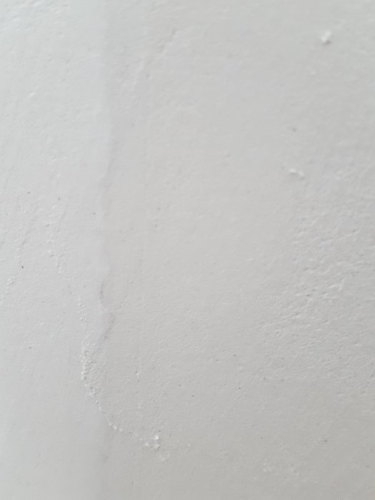 Något ojämn vit gipsvägg med en reparerad, spacklad spricka som sträcker sig vertikalt.