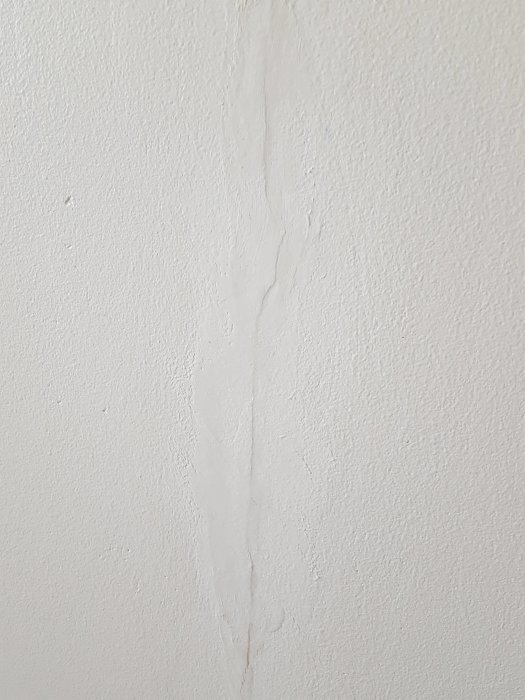 Nytätad djup spricka i vitmålad gipsvägg, delvis ojämnt spacklad.