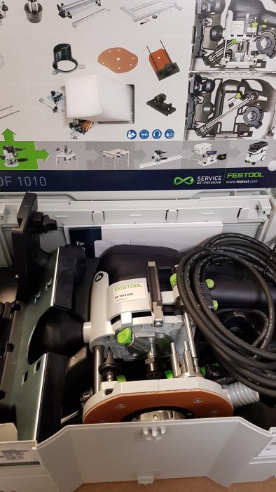 Festool OF 1010 EBQ-set i en öppnad verktygslåda med fräs och tillbehör synliga mot en illustrerad produktkatalog.