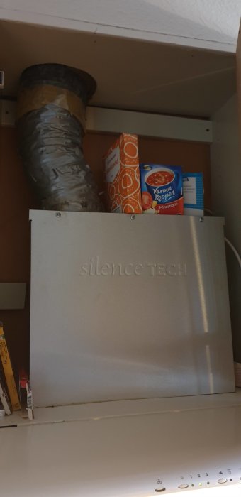 Gammal underbyggd köksfläkt med slitna knappar under skåp och ventilationsrör, omgiven av förpackningar.