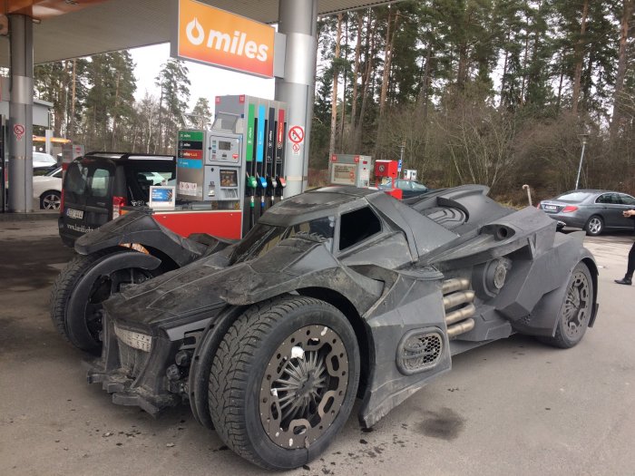 En Batmobile-liknande bil tankar vid en bensinstation med andra fordon och skog i bakgrunden.