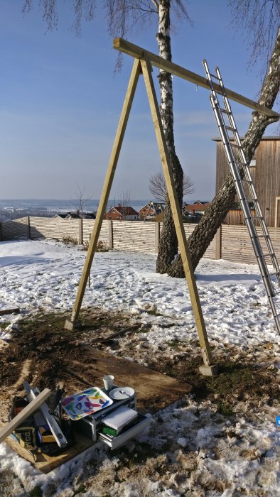 Nybyggd gungställning av trä med infästning i ett träd, verktyg och material på marken täckt av snö.