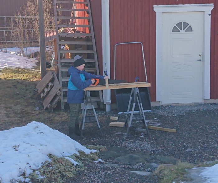 Barn sågar trä med fogsvans vid arbetsbänk utomhus nära röd stuga.