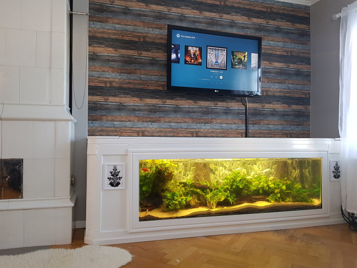 Vardagsrum med väggmonterad TV och inbyggt akvarium i vit möbel längs väggen.