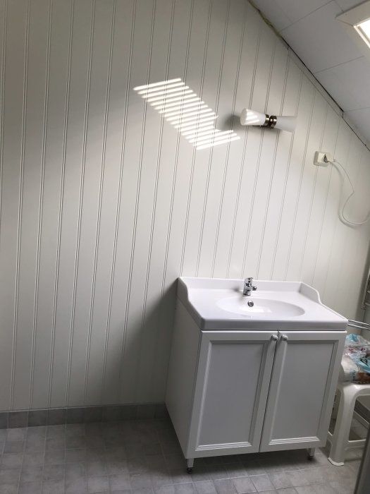 Ett vitt skåp med handfat i ett ljust badrum med väggpanel och grå klinkergolv, färdigställande återstår.
