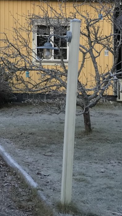 Stolpe med belysningsarmatur fastskruvad, reflektion syns, mot gult hus och bar trädgren i förgrunden.