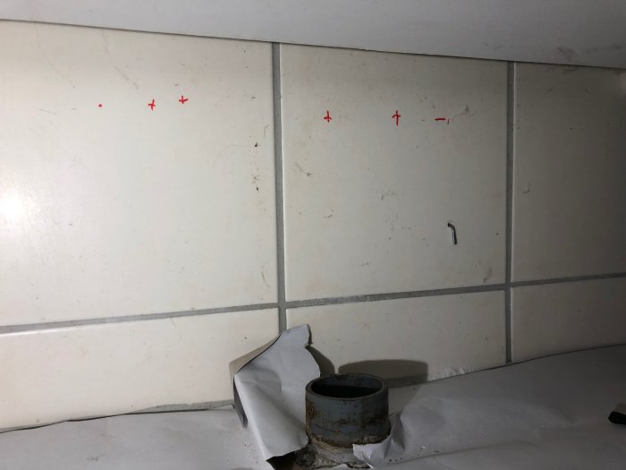 Avloppsrör som sticker upp ur betonggolv vid vägg med röda kryssmarkeringar.