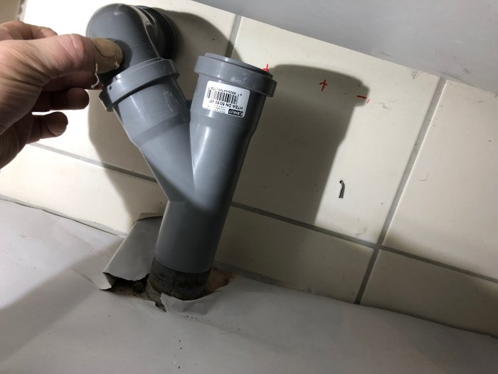 PVC-avloppsrör som kommer upp ur en gjuten platta, med två 90-gradersböjar och röda kryss markerade på en kakelvägg.