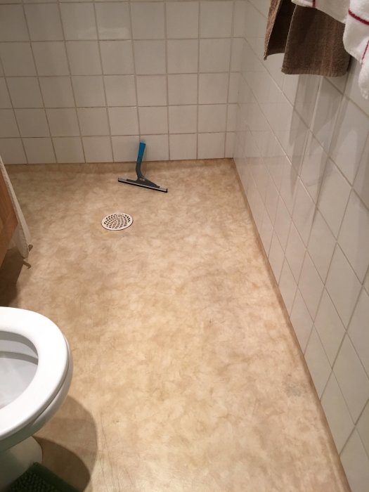 Oklart badrumsgolv med en golvskrapa, vita kakelväggar och en toalettstol synlig till vänster.