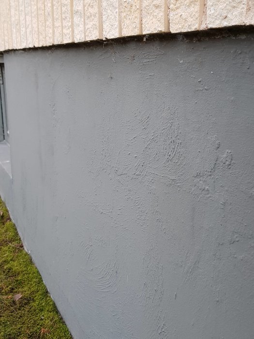 Målade grå grundmuren på ett hus med synliga lagningar och lätt ojämn yta, intill grönt gräs.