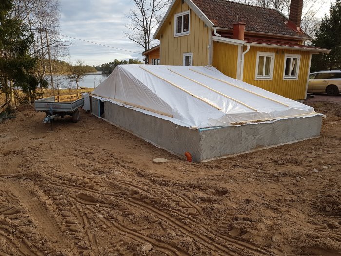 Grund till utbyggnad med dagvattenrör synligt, täckt med presenning, intill ett gult hus.