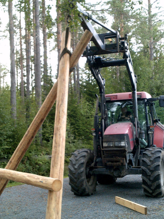 Traktor med lyftanordning hanterar en stor träbjälke vid skogskant.