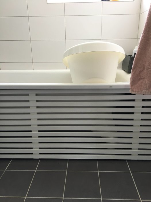 Vit badrumshink i ett badkar med vit frontpanel och mörkgråa kakelplattor.