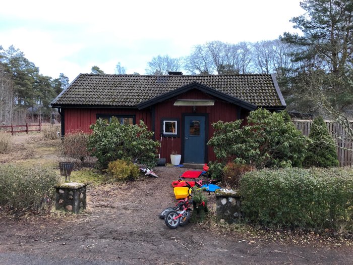 Rött hus med svart tegeltak och leksaker på en rörig baksida, omgivet av grönska och staket.