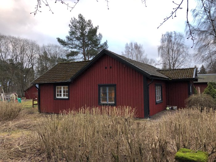 Rött trähus med flera takvinklar, omgivet av träd och en lekplats syns i bakgrunden.