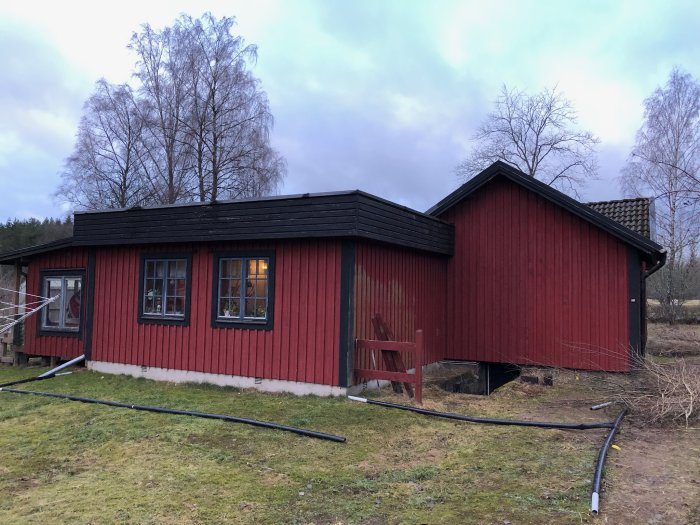 Rödmålat hus med svart tak och komplex takkonstruktion, visar baksidan med rör och bråte på marken.