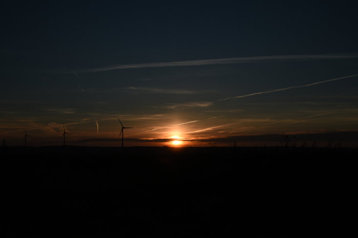 Solnedgång över ett landskap med vindkraftverk mot en dramatisk himmel.