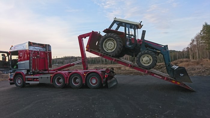 Ny traktor lastas av från transportlastbil på en landsväg, skog i bakgrunden.