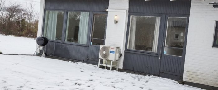Delvis täckt av snö husfasad med blå panel och vita fönsterkarmar, värmepump till höger.
