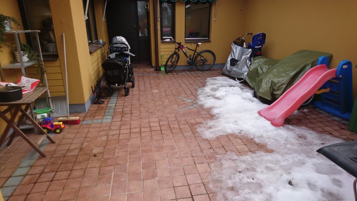 Delvis snötäckt uteplats med rödaktiga italienska plattor och leksaker, barnvagn och cykel längs väggen.