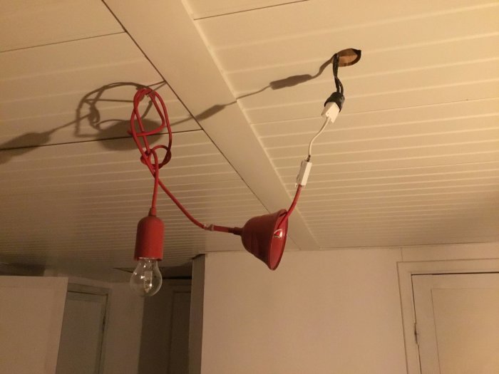 Improviserad taklampa med glödlampa hängande från röd sladd vid ett spartanskt boende.