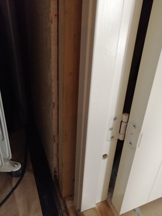 En snedställd dörrkarm monterad bredvid ett stycke ojämnt trä för att hålla karmen rak, med synliga gångjärn.