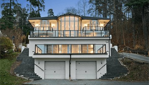 Modernt hus med två våningar och vita fasader, beläget på en kulle med garage i bottenvåningen.