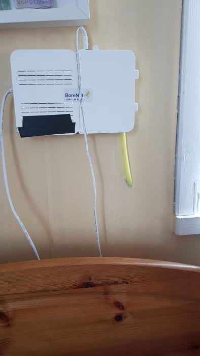 En vit router med svart tejp på och synliga kablar längs en vägg invid ett fönsterfoder.