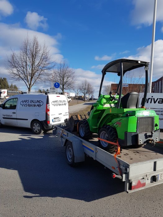 Grön grävmaskin fastspänd på en släpvagn parkerad på en gata.