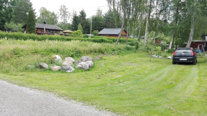 Ojämn gräsyta med spridda stenblock framför en häck, bredvid en grusväg och en parkerad bil, med stugor i bakgrunden.