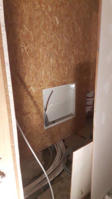 Förrådsutrymme under trapp med öppen elcentral och rullar av vinylgolv.