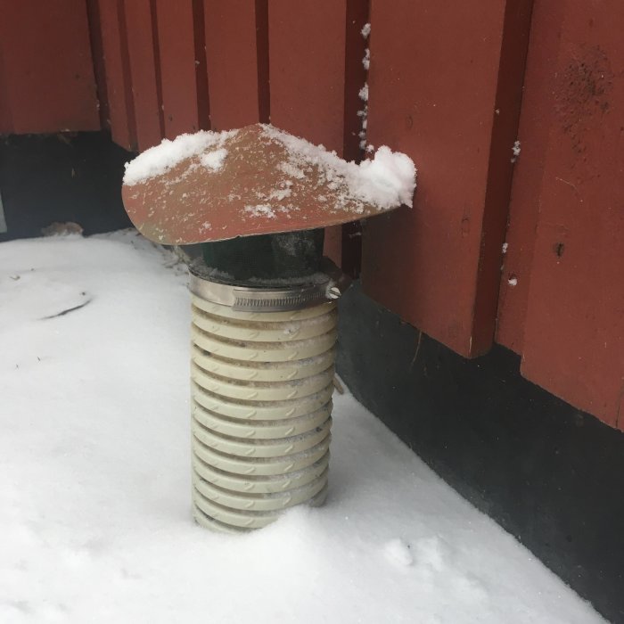 Ett rör med en hattformad topp i snö intill röd vägg, eventuellt del av äldre avfuktningssystem.