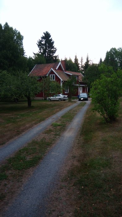 Rödmålat trähus med vita knutar och tegeltak, omgivet av grönska och en grusväg som leder fram till huset.