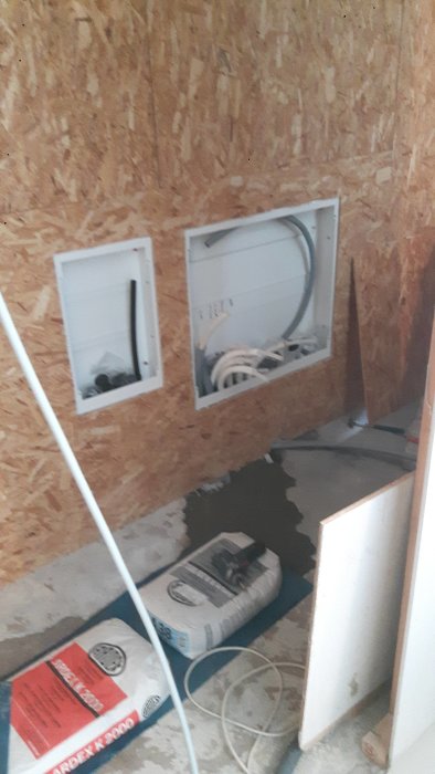 Installationsvägg med öppet tappvattenskåp, säckar som tyngd och osynlig fönsterbänk.