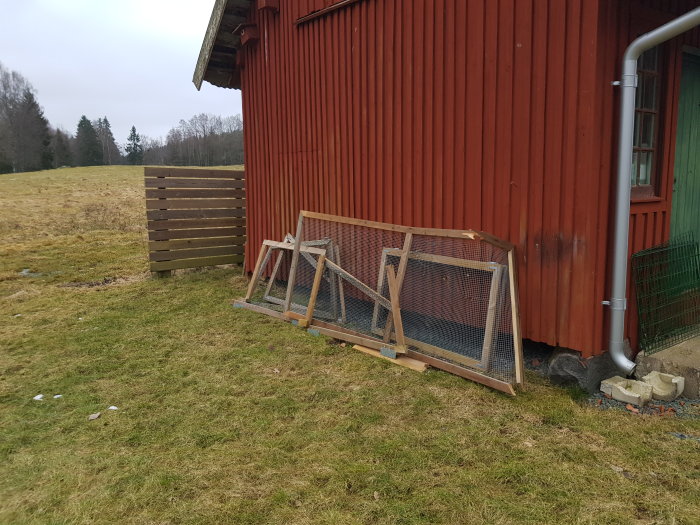 Träramar med nät står lutade mot en röd stuga vid sidan av en gräsmatta.