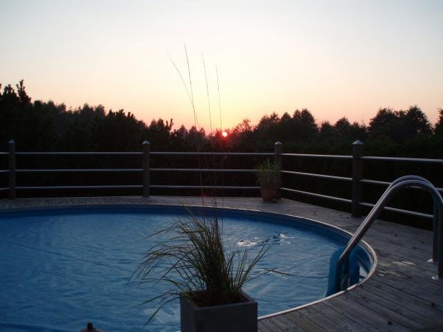 Solnedgång bakom träd från en terrass med en rund pool och trappräcke.