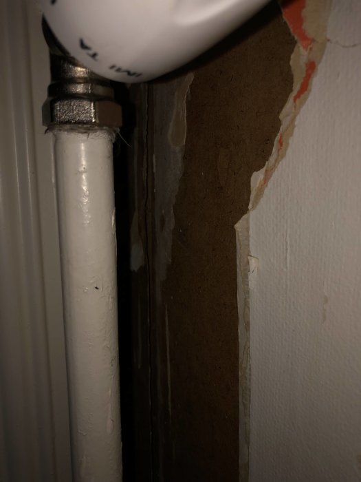 Skadad masonitskiva med sprickor bakom ett vitt radiatorrör i ett äldre hus.