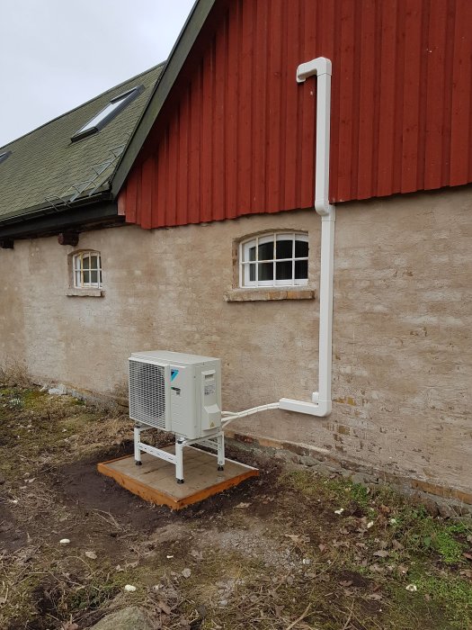 Luftvärmepump installerad utanför en tegelvägg med rörledningar upp längs en rödfärgad trävägg.