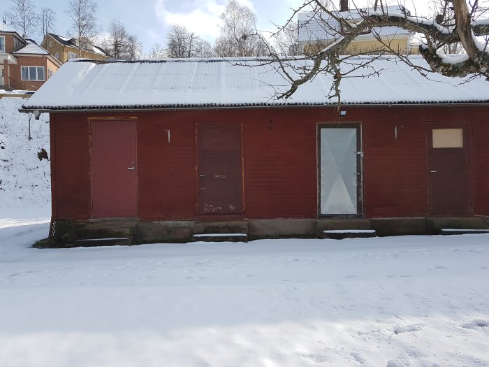 Röd träbyggnad som potentiellt är ett gammalt garage eller lada med snötäckt tak, i ett vinterlandskap.