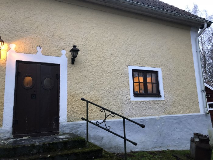 Exteriör av ett gammalt stenhus med träporthusdörrar och belyst fönster i skymning.
