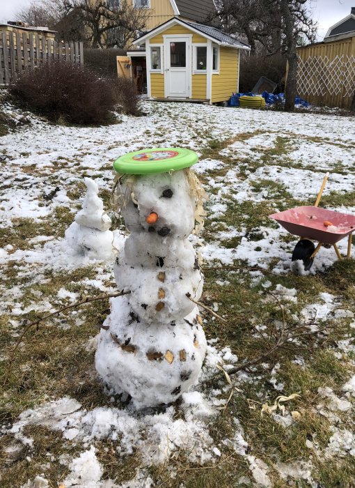 En hemmagjord snögubbe med en grön leksak som hatt och en morot som näsa i en trädgård med lite snö.