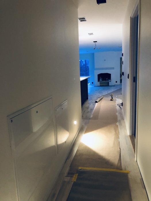 Korridor under renovering med inbyggda väggskåp, skyddsplast på golvet och synlig öppen spis i bakgrunden.