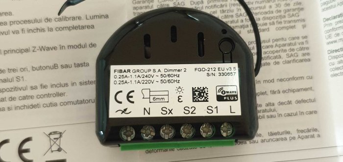 En Fibaro Dimmer 2-enhet med textetikett, anslutningsterminaler och en ansluten svart kabel, mot en textbakgrund.