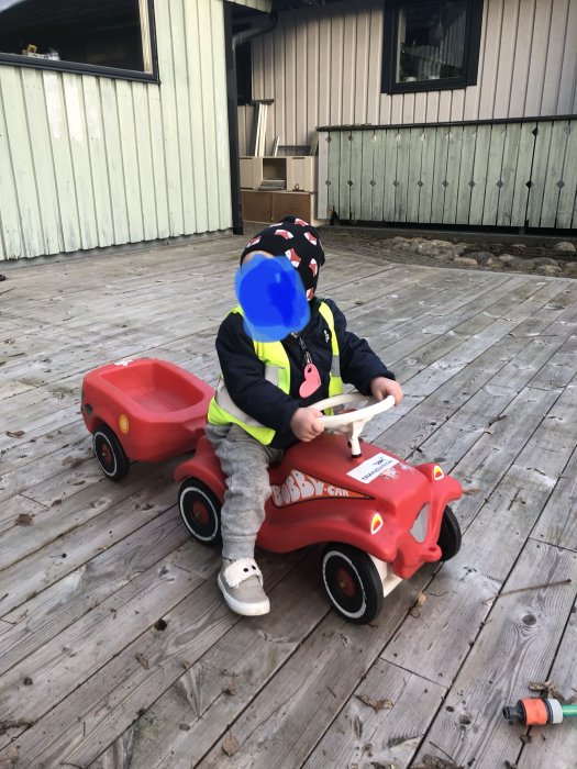 Barn leker med en röd Bobby-Car leksaksbil och släpvagn på en träveranda.