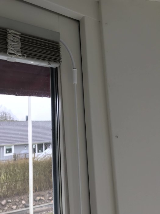 Nytt vred installerat på en vit persienn i ett fönster, med utsikt mot ett bostadsområde.