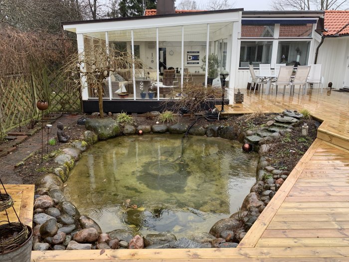 Trädgårdsdamm omgiven av stenar och vinterföna växter med en tillhörande altan och uteplats.