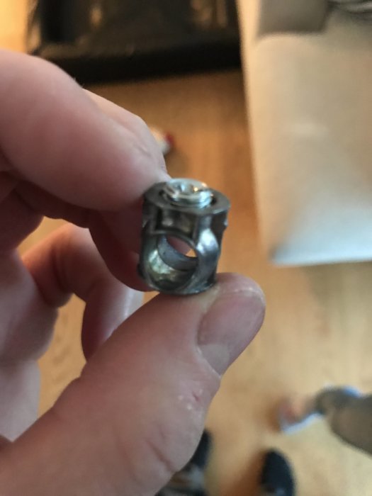 Hand håller en metallmutter på cirka 15mm mot suddig inomhusbakgrund.