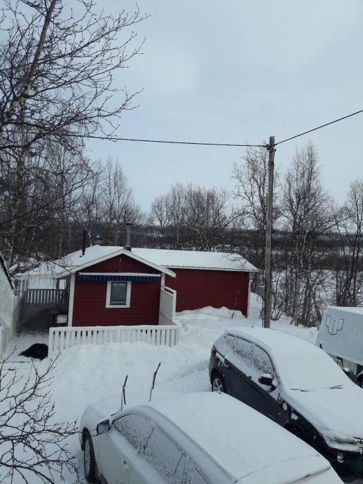 Röd stuga med snötäckta bilar framför, vinterlandskap.