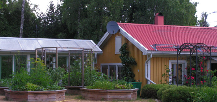 Översikt över en trädgård med odlingsbäddar, växthus och ett gulmålat hus med rött tak.